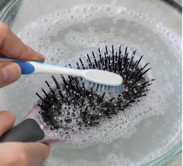 Small cleaning brush scrubbing between the bristles of a wet paddle hairbrush