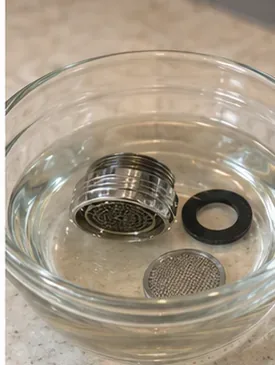 A faucet aerator, screen, and rubber gasket soaking in a clear bowl to loosen mineral deposits.