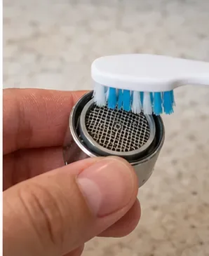 A small soft toothbrush gently scrubbing the mesh of a removed faucet aerator.