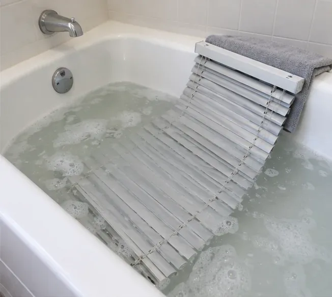 Gray horizontal blind slats resting in lukewarm soapy bathwater while the white headrail stays on a towel above the waterline.