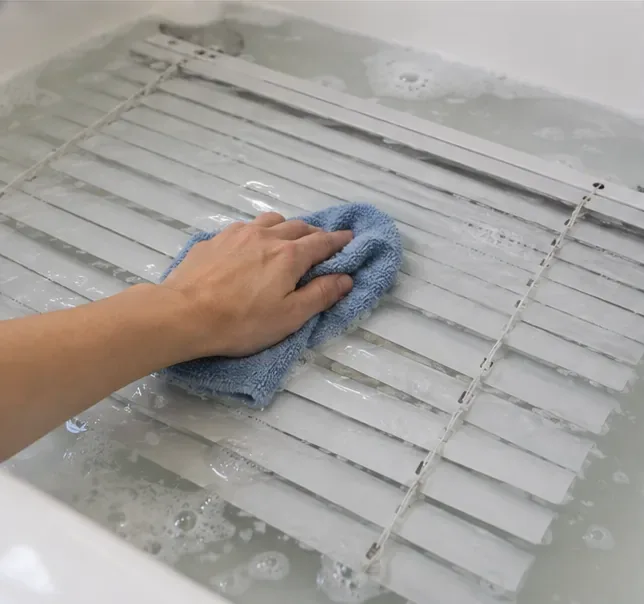 Hand wiping wet gray blind slats with a blue microfiber cloth in a bathtub.