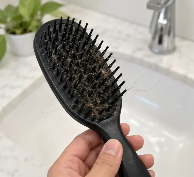 A hand holds a black hairbrush with visible trapped hair and buildup before cleaning.