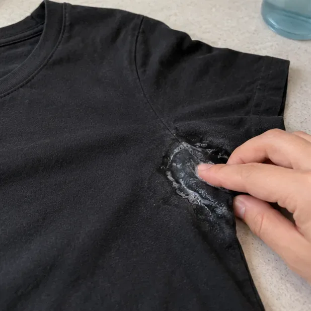 A small amount of liquid cleaner being gently worked into deodorant buildup at the underarm of a black T-shirt.