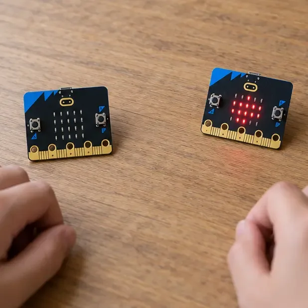 Kaksi micro:bit-korttia pöydällä, toisessa punainen LED-kuvio ja kädet korttien lähellä